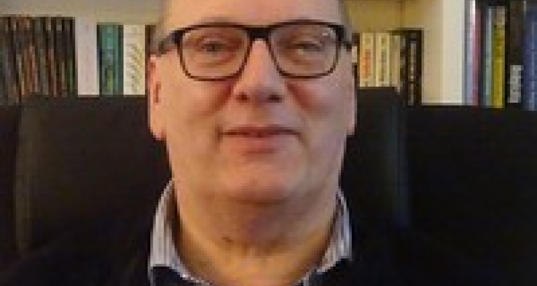 Een man met een bril zit in een stoel voor een boekenplank.