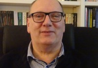 Een man met een bril zit in een stoel voor een boekenplank.