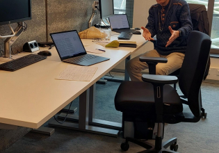 Een man zit aan een bureau met twee computers en documenten.