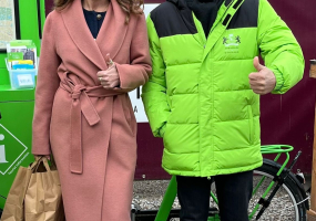 Een vrouw in een roze jas en een man in een groene jas staan met een duim omhoog.