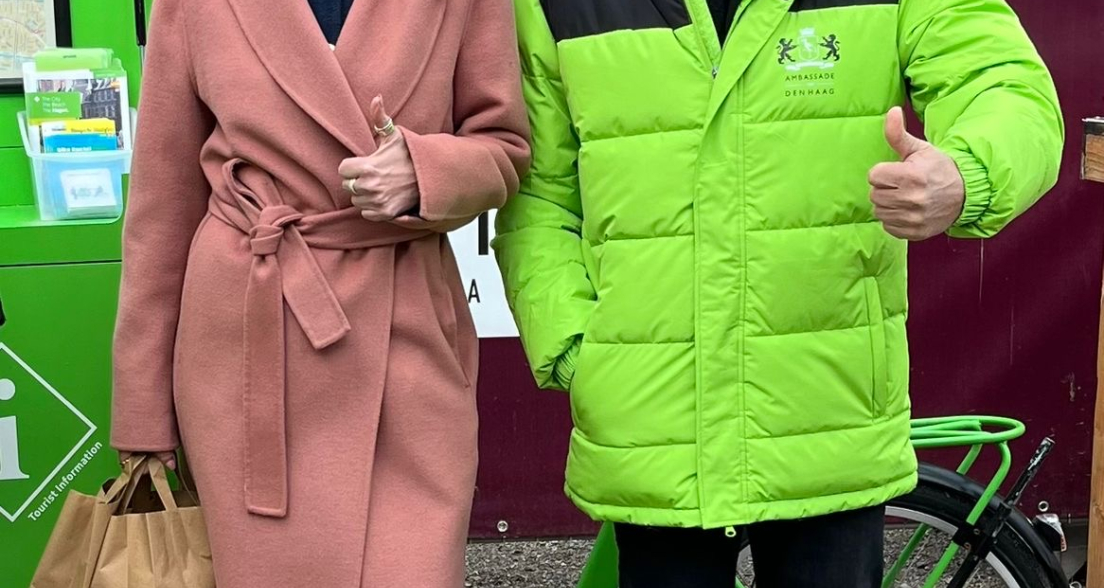 Een vrouw in een roze jas en een man in een groene jas staan met een duim omhoog.