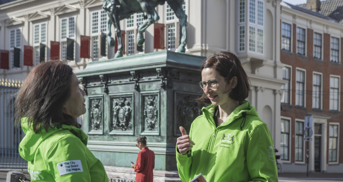 Twee vrouwen in groene jassen praten bij een standbeeld in Den Haag.