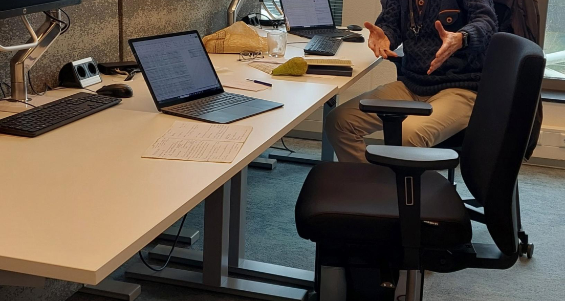 Een man zit aan een bureau met twee computers en documenten.